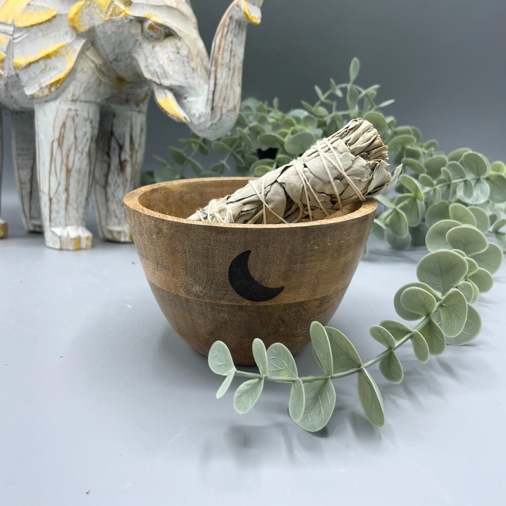Wooden bowl with sage inside, surrounded by eucalyptus leaves and a stone elephant figurine.