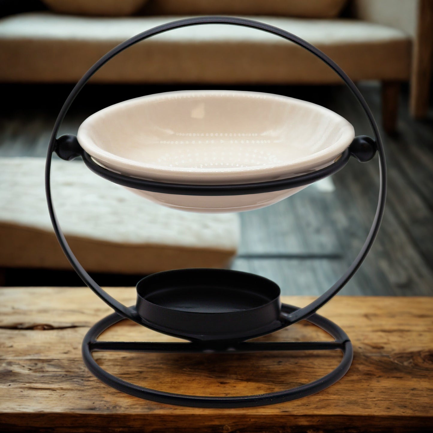 Lifestyle photo of black circular metal oil burner with cream ceramic dish on a rustic wooden table; tealight tray below.