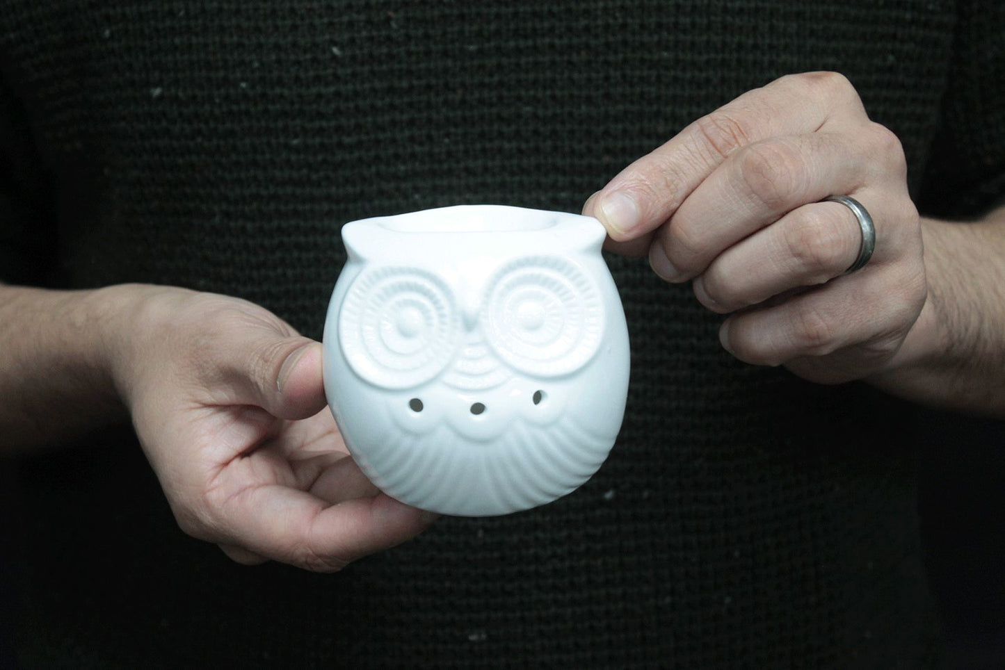 lose-up of a glossy white ceramic oil burner shaped like an owl, with round carved eyes and feather details, held gently in two hands against a dark knitted jumper.