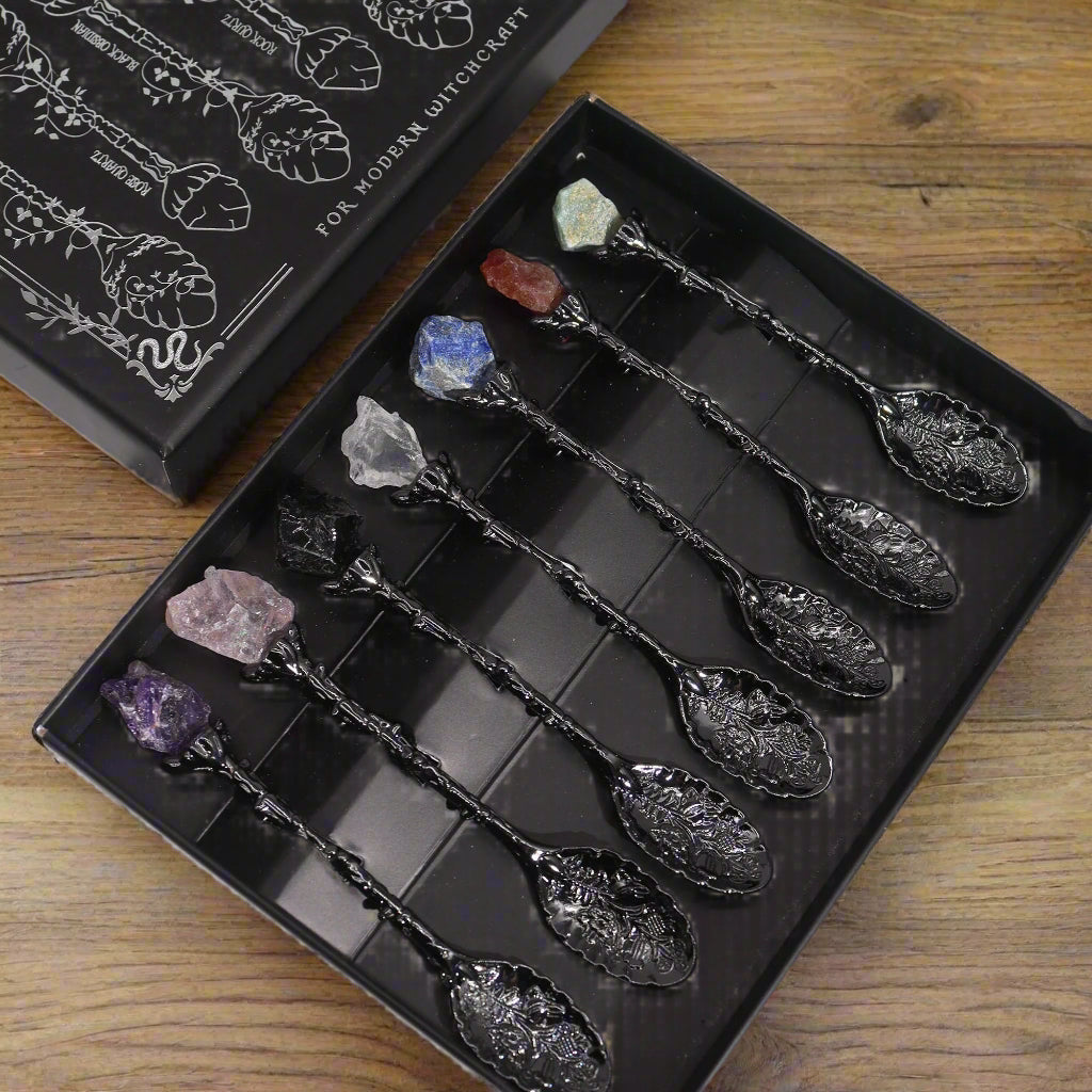 Open gift box showing seven dark-silver teaspoons topped with rough-cut gemstones arranged in a row.