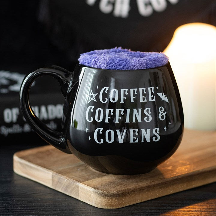Black mug with 'Coffee Coffins & Covens' text on a wooden coaster.