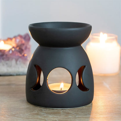 Black ceramic oil burner with moon phases on a wooden surface, surrounded by candles.