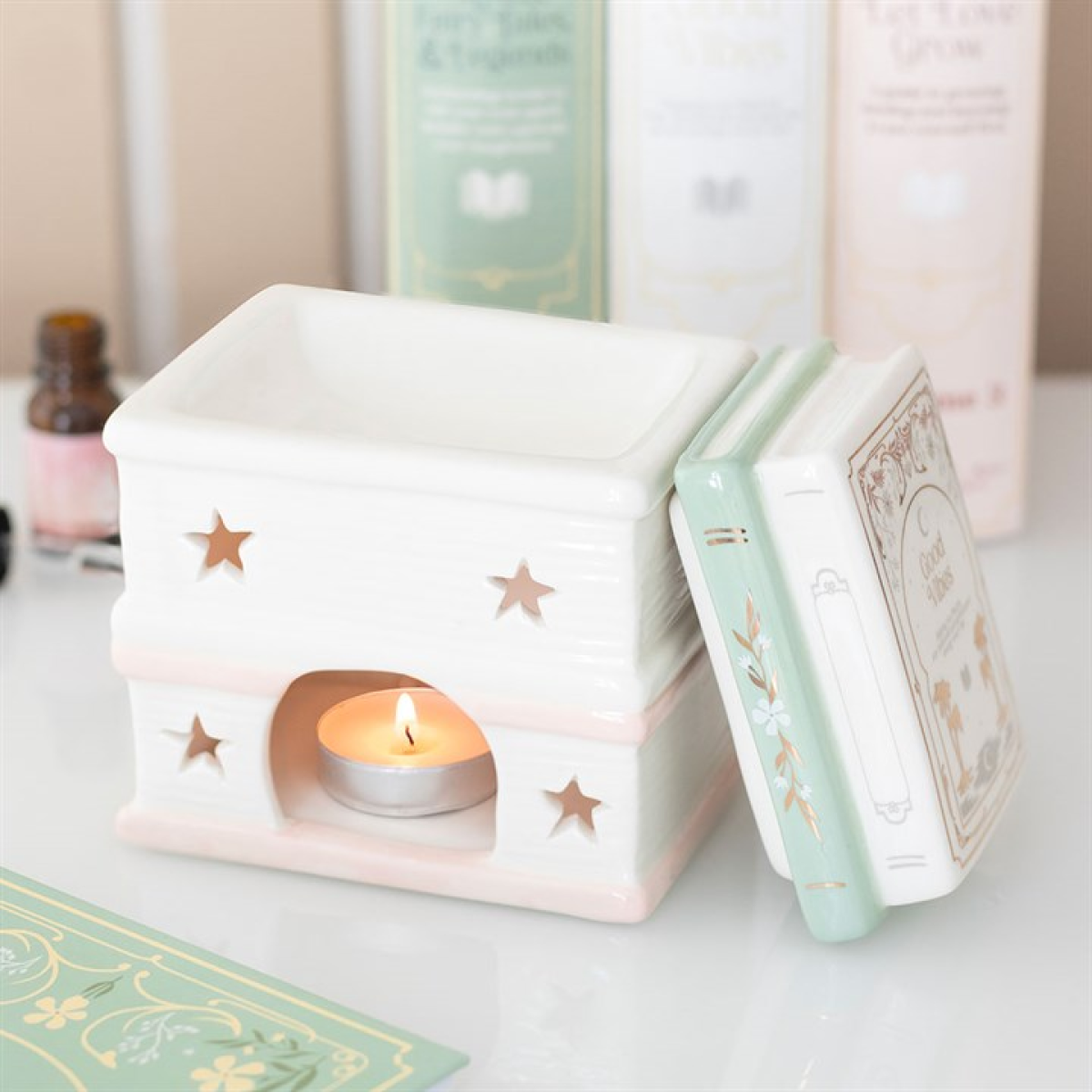 Ceramic oil burner shaped like stacked books in pastel sage and blush with star cut-outs; tealight glows beneath the removable-lid dish. Fae Cottage