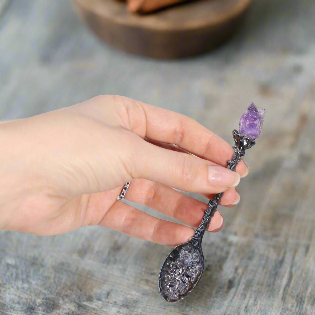 Hand holding a decorative spoon with a Amethyst crystal on a dark grey spoon 