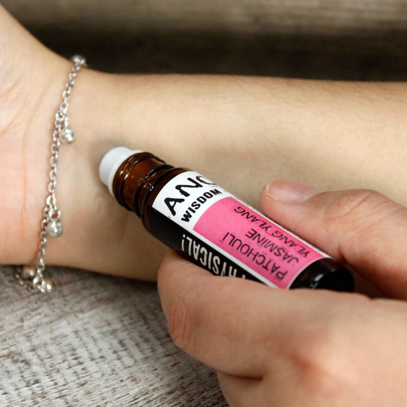 Small amber glass roller bottle with black cap and stainless-steel ball, labelled ‘Get Physical! Patchouli • Jasmine • Ylang Ylang,’ shown being rolled onto the inside of a wrist for pulse-point application