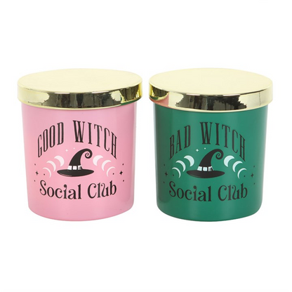 Two scented glass candles, one pink “Good Witch” and one green “Bad Witch,” lit and displayed with gold lids; giftable set.