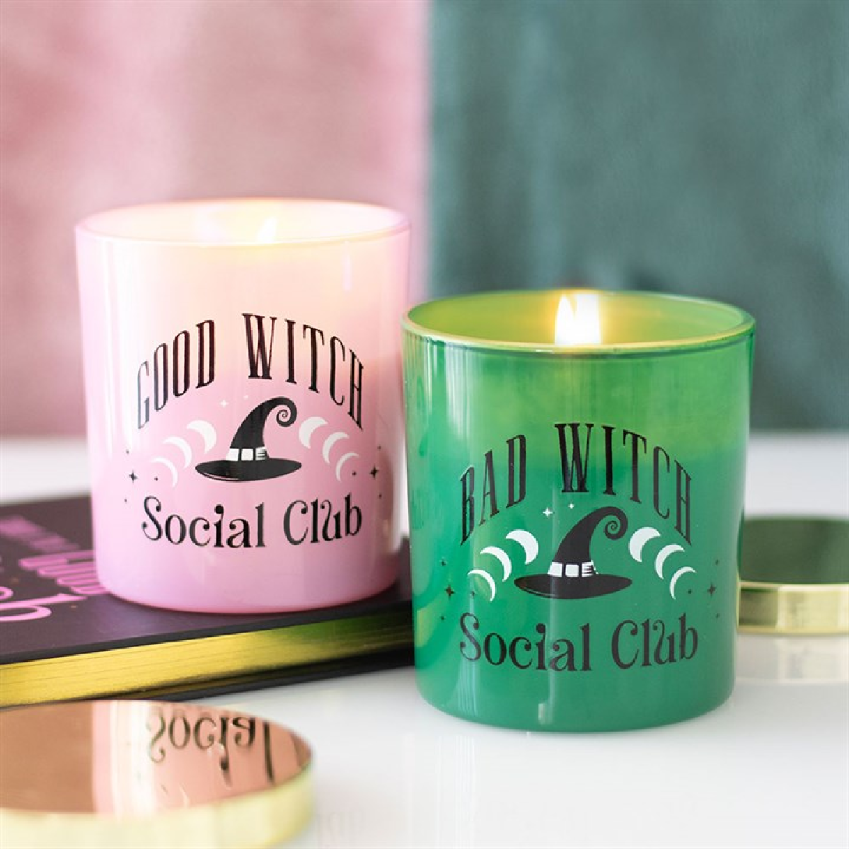 Two scented glass candles, one pink “Good Witch” and one green “Bad Witch,” lit and displayed with gold lids; giftable set.