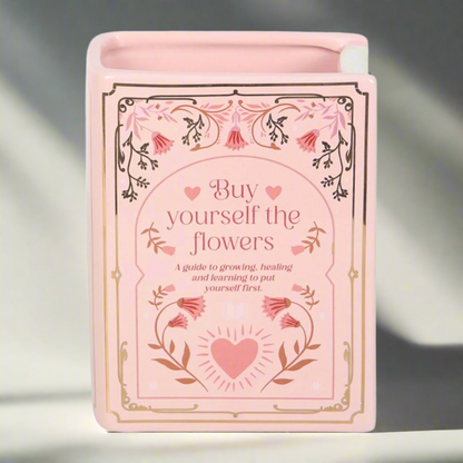 Pink ceramic vase shaped like an upright book with gold floral detailing and the phrase ‘Buy yourself the flowers,’ 