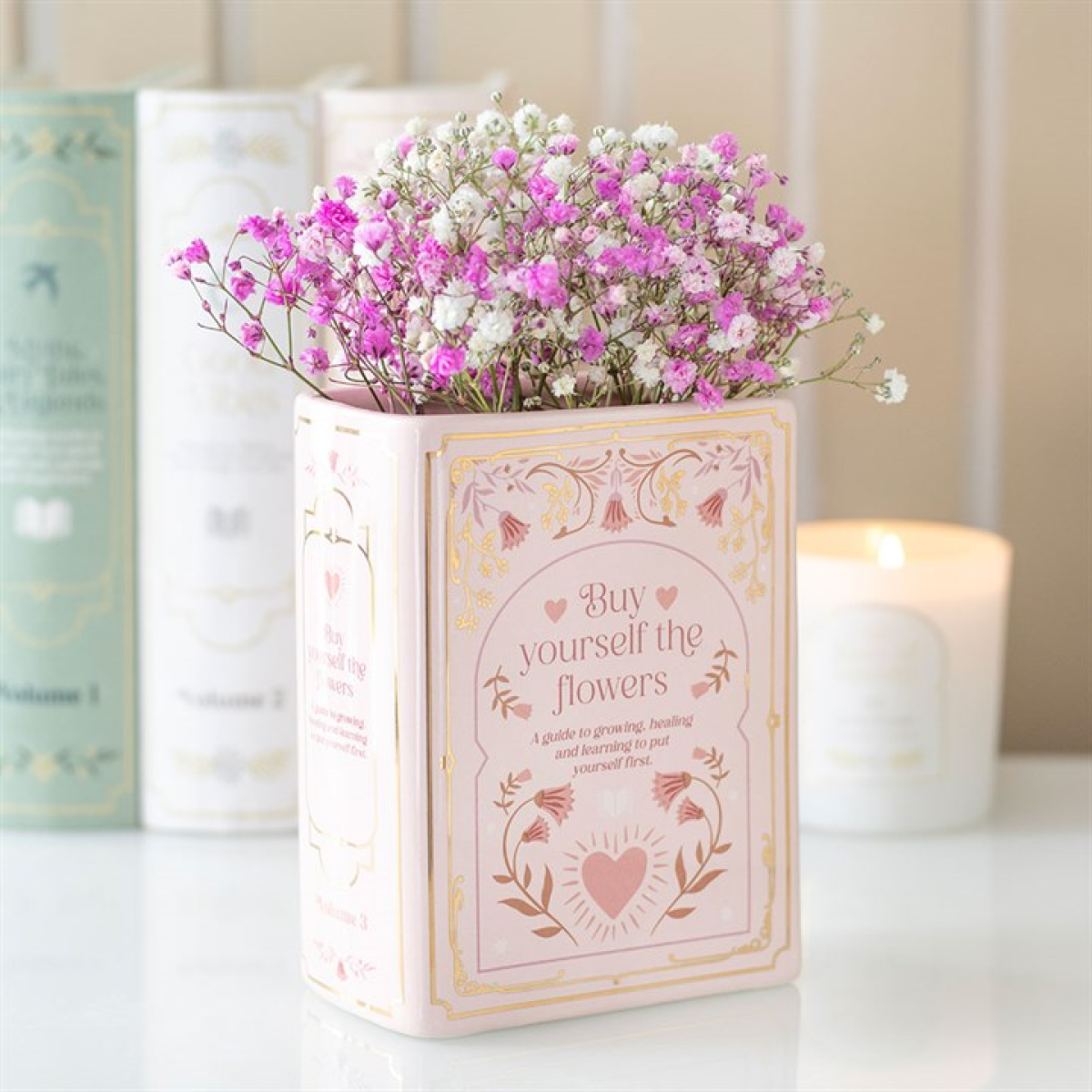 Pink ceramic vase shaped like an upright book with gold floral detailing and the phrase ‘Buy yourself the flowers,’ styled with small pink and white blooms.