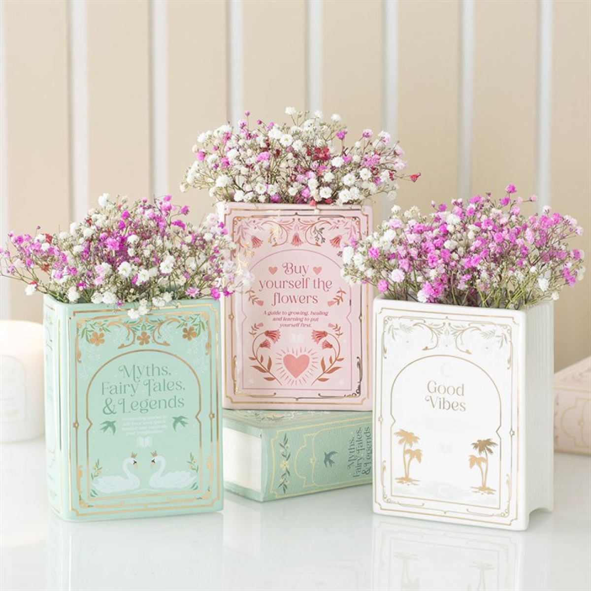 trio of faux book vases in sage green 'Myths. Fairy Tales, & Legends', pale pink ‘Buy yourself the flowers,’ and white & gold 'Good Vibes' styled with small pink and white blooms. 