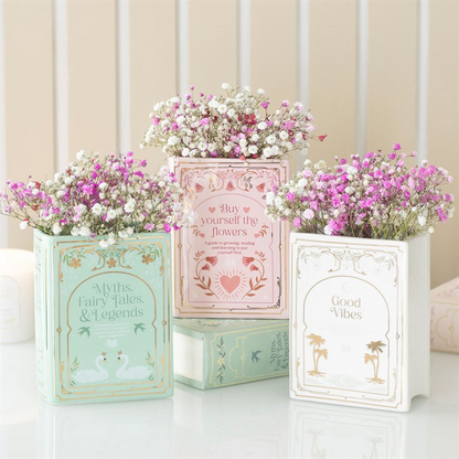 trio of faux book vases in sage green 'Myths. Fairy Tales, & Legends', pale pink ‘Buy yourself the flowers,’ and white & gold 'Good Vibes' styled with small pink and white blooms. 