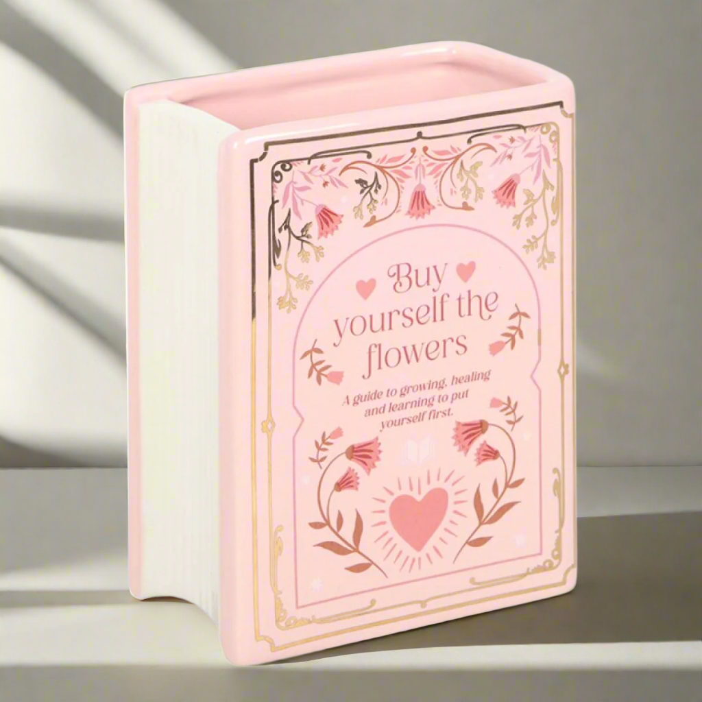 Pink ceramic vase shaped like an upright book with gold floral detailing and the phrase ‘Buy yourself the flowers,’ 